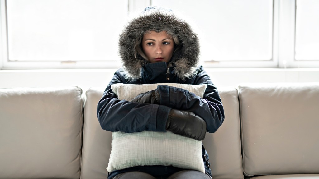 Eine Frau sitzt vollständig mit Jacke angezogen in ihrer Wohnung.