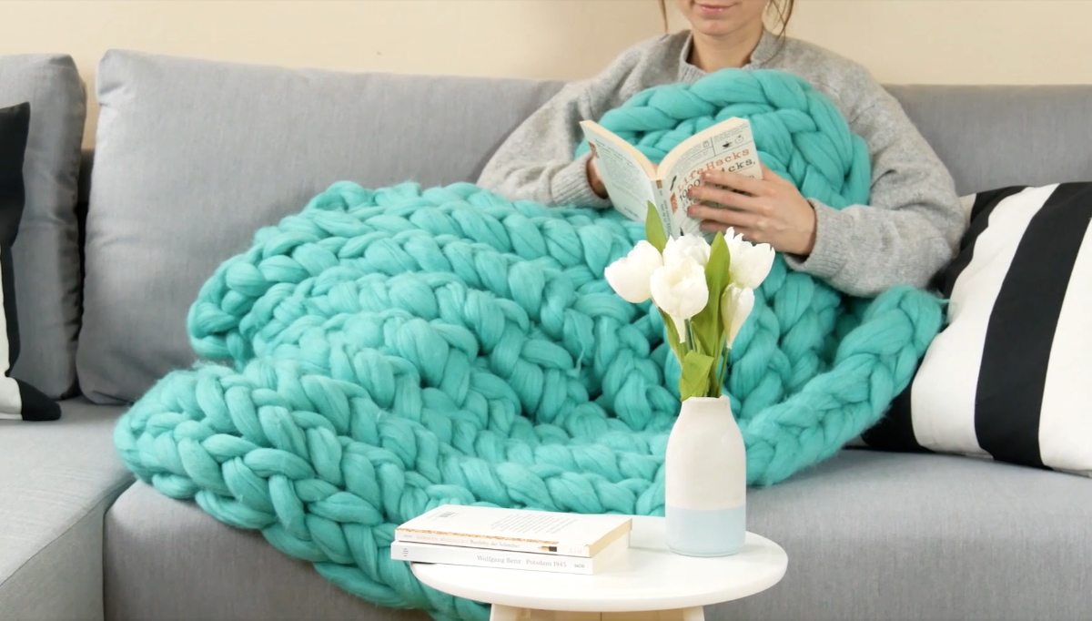 Eine junge Frau liegt mit einer selbstgestrickten Decke auf einer Couch und liest ein Buch.
