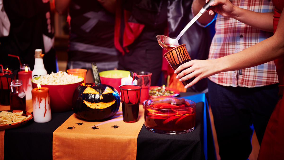 Ein Buffet auf einer Halloween-Party. Man sieht gruselige Deko und eine Frau gießt sich Bowle in einen Becher.