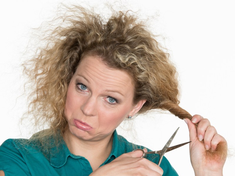 Eine Frau mit Locken vor weißem Hintergrund. Sie hat eine Strähne in der Hand, an die sie eine Schere hält.