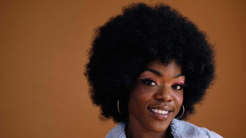 Eine Schwarze Frau mit Afro vor einem hellbraunen Hintergrund.