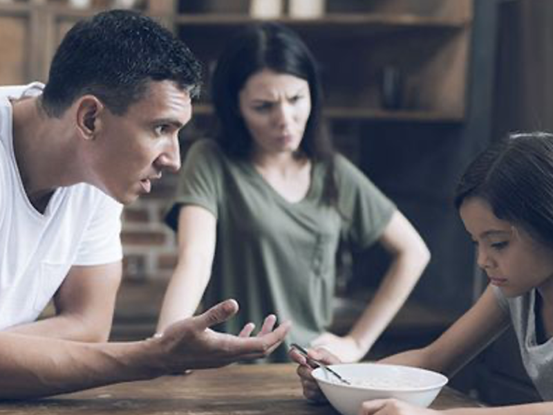 Ein heteronormatives Paar schimpft mit seiner Tochter. Das Mädchen schaut traurig auf ihre Müslischüssel