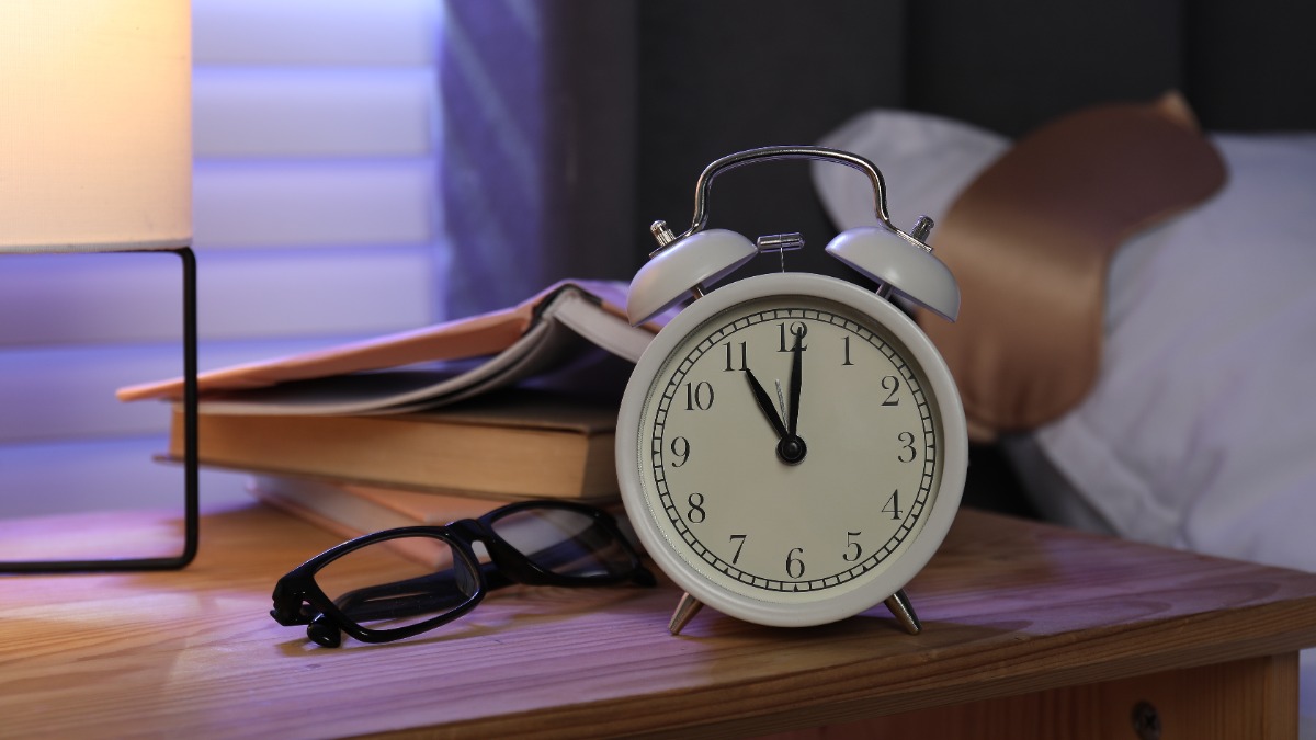 ein Wecker, eine Brille und ein Buch neben einem Bett