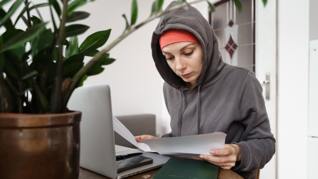 Eine Frau sitzt am Schreibtisch und schaut sich Papiere an. Sie trägt eine Mütze und darüber hat sie die Kapuze ihres Hoodies gezogen.