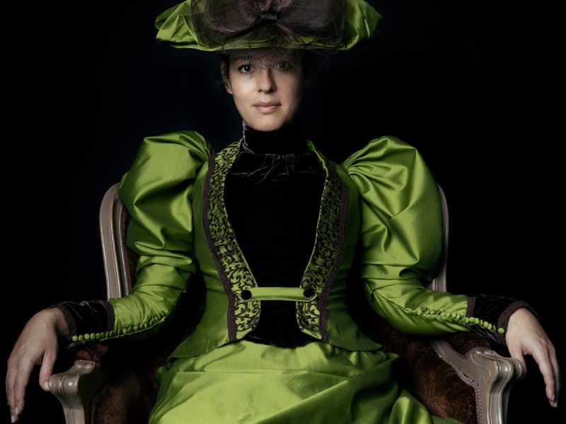 Eine Frau in einem giftgrünen viktorianischen Kleid sitzt auf einem Stuhl.