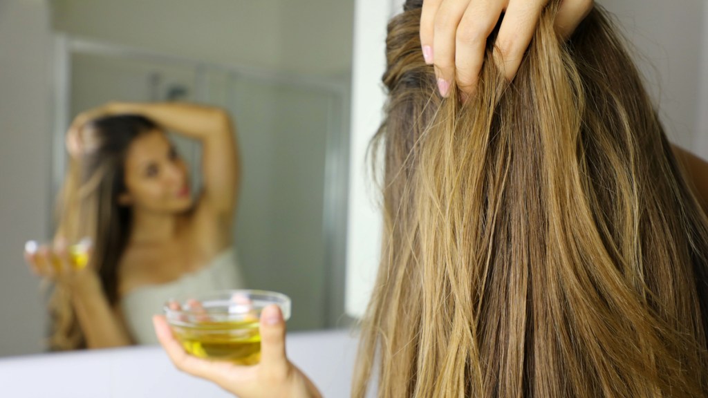 Eine Frau reibt Öl in ihre Haare, während sie sich im Spiegel anschaut.