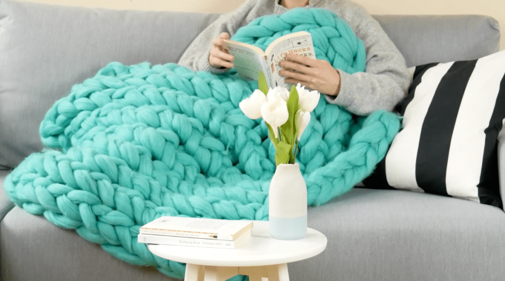 Eine Frau liegt mit einer Wolldecke auf einem Sofa und liest.