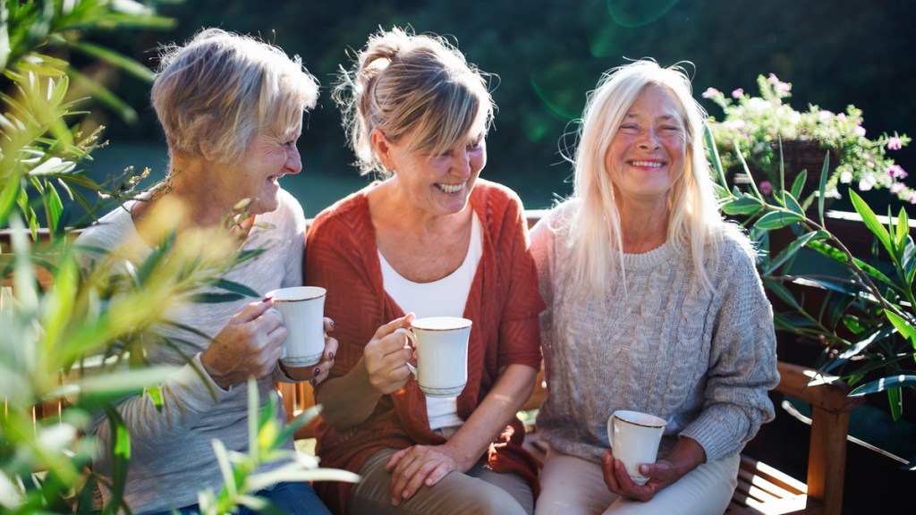 Drei Frauen mittleren Alters sitzen draußen auf einer Bank und lachen.