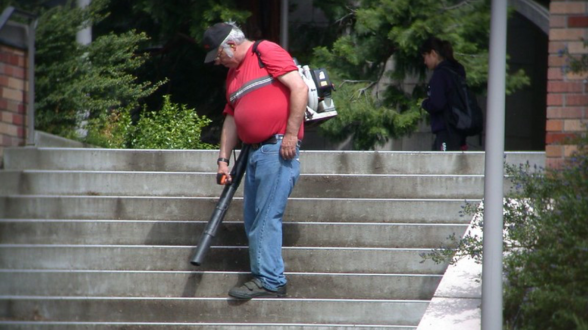 Ein älterer Mann in rotem Shirt und Jeans pustet eine Treppe, auf der keine Blätter liegen, mit einem Laubbläser ab.