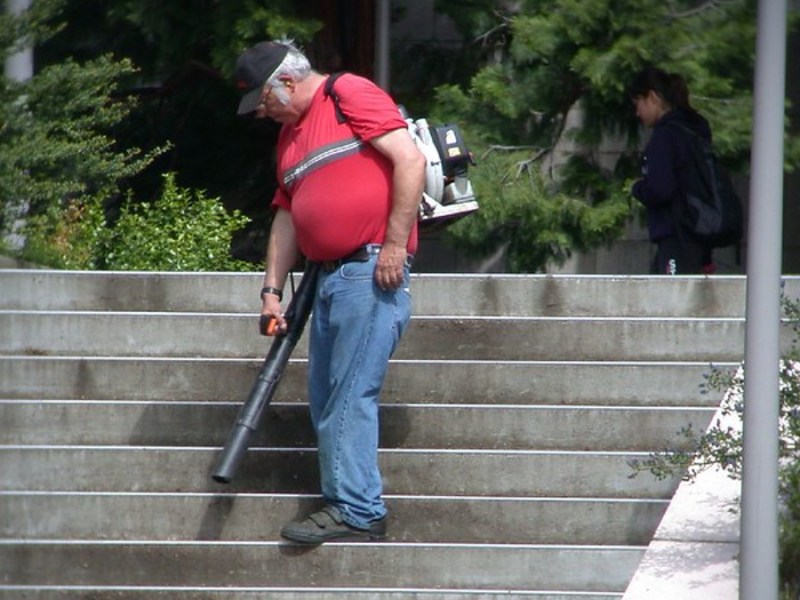Ein älterer Mann in rotem Shirt und Jeans pustet eine Treppe, auf der keine Blätter liegen, mit einem Laubbläser ab.