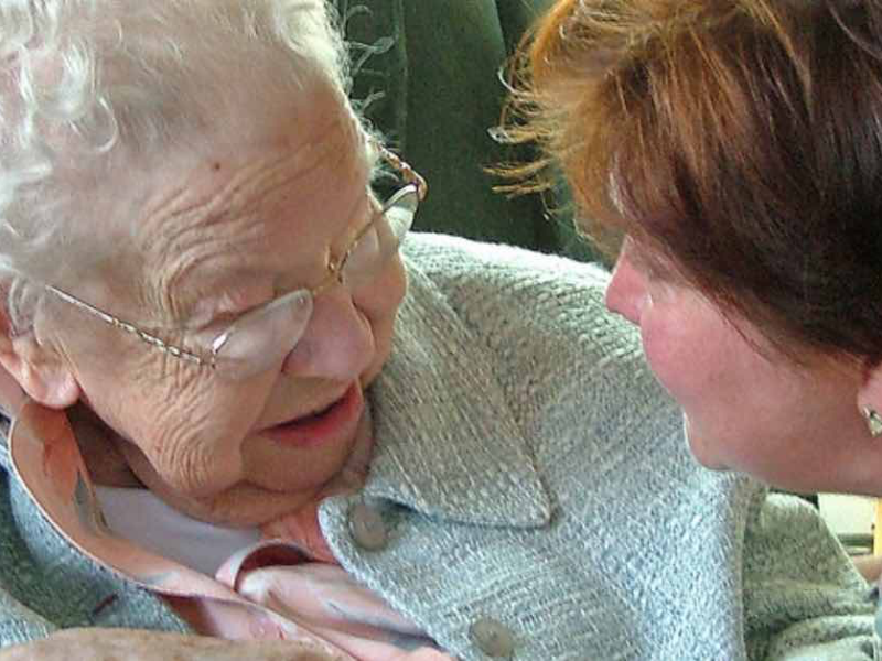 Demenz vorbeugen: Frau spricht freundlich mit Seniorin und hält ihre Hand.