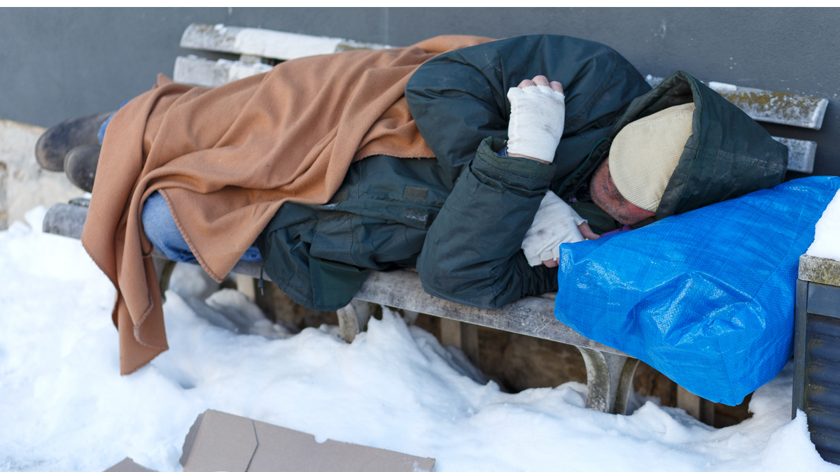 Ein obdachloser Mann schläft mit Decken bedeckt auf einer Bank im Winter.