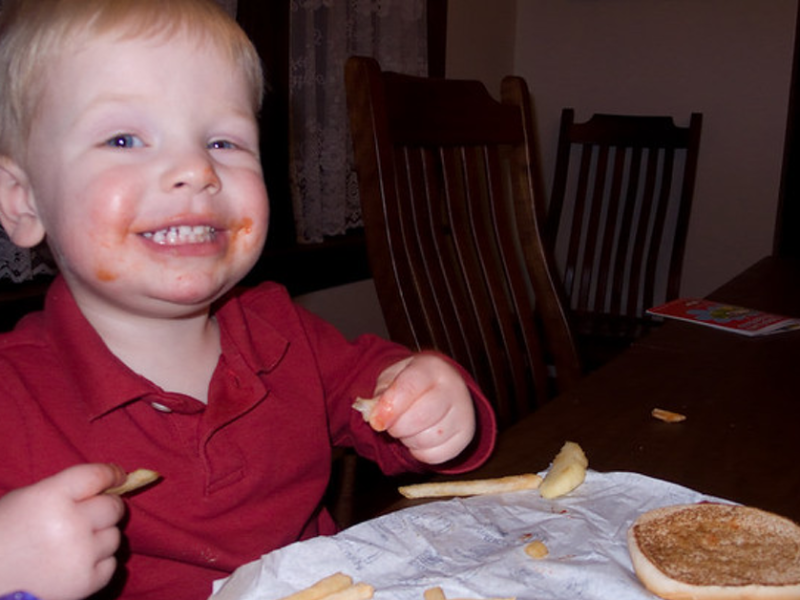 Kleinkind sitzt am Tisch, ist im Gesicht mit Essen verschmiert und grinst in die Kamera