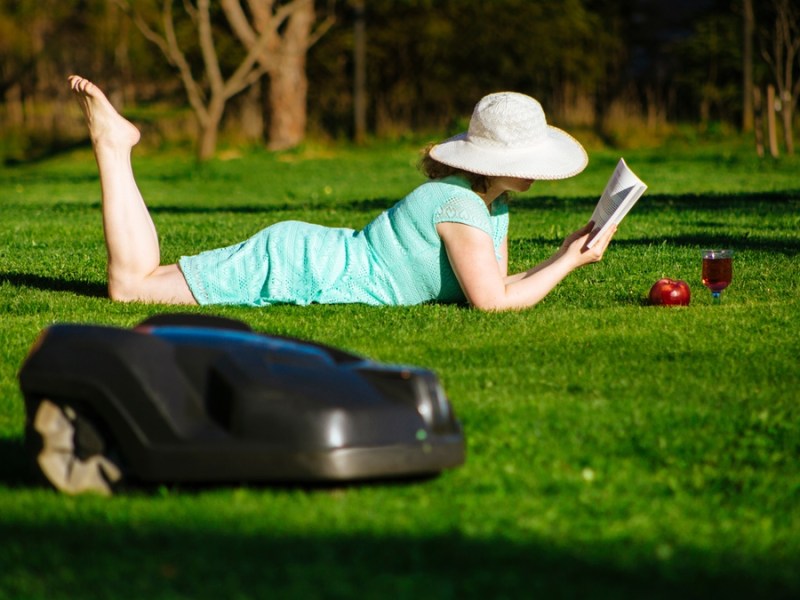 Eine Frau liest ein Buch auf ihrem Rasen, auf dem ein Mähroboter mäht.