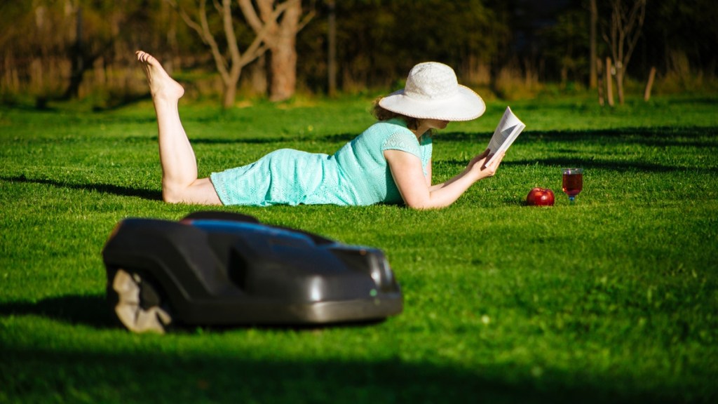 Eine Frau liest ein Buch auf ihrem Rasen, auf dem ein Mähroboter mäht.