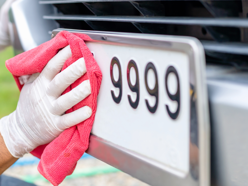 Eine Person säubert ein Nummerschild, das mit 9999 endet, mit einem roten Tuch. Dabei trägt die Person weiße Einmalhandschuhe.