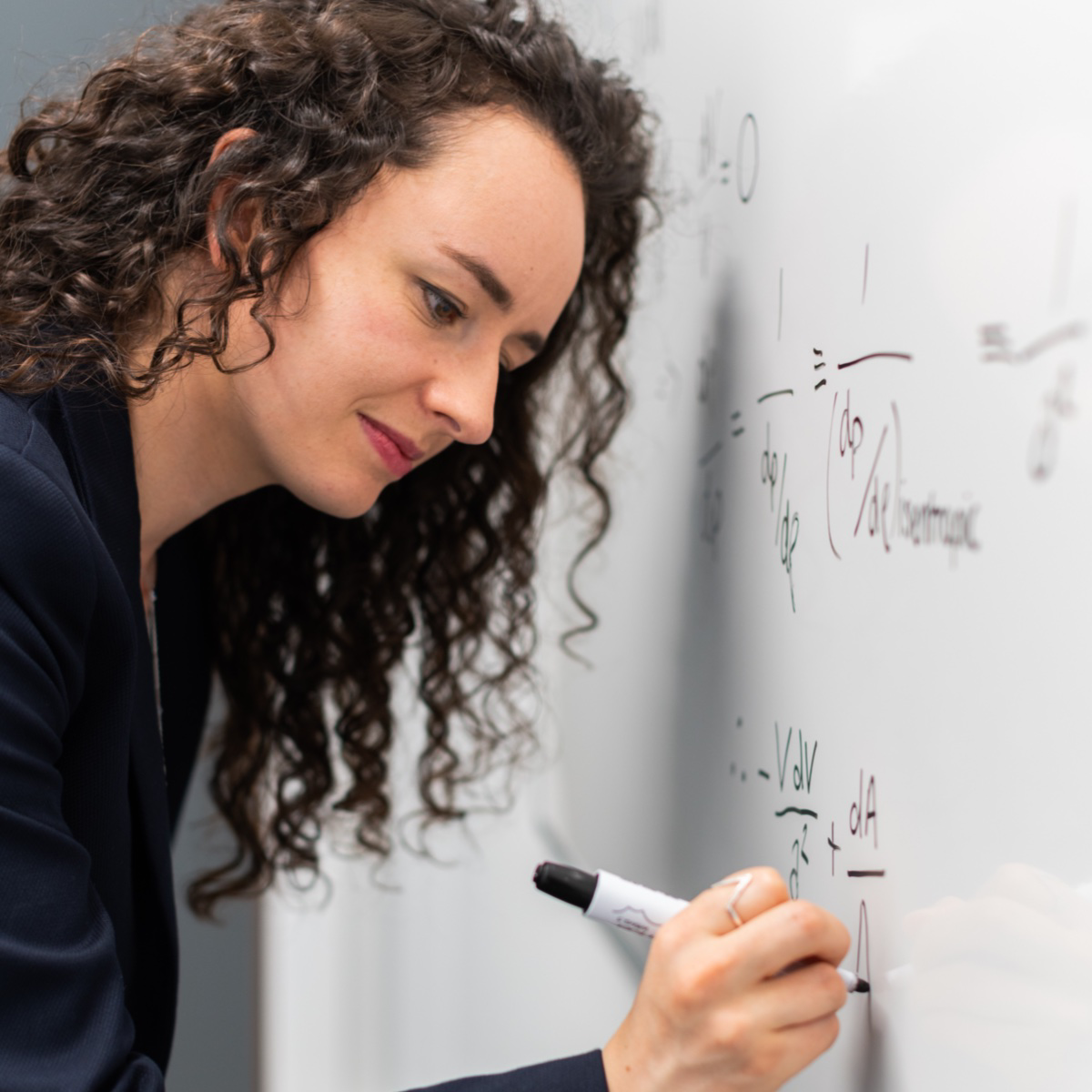 Schick gekleidete Frau schreibt mathematische Formeln auf eine weiße Tafel.