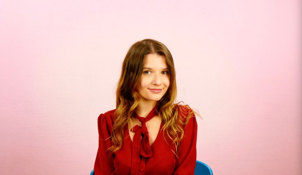 Eine Frau mit welligen Haaren sitzt vor einer pinken Wand.