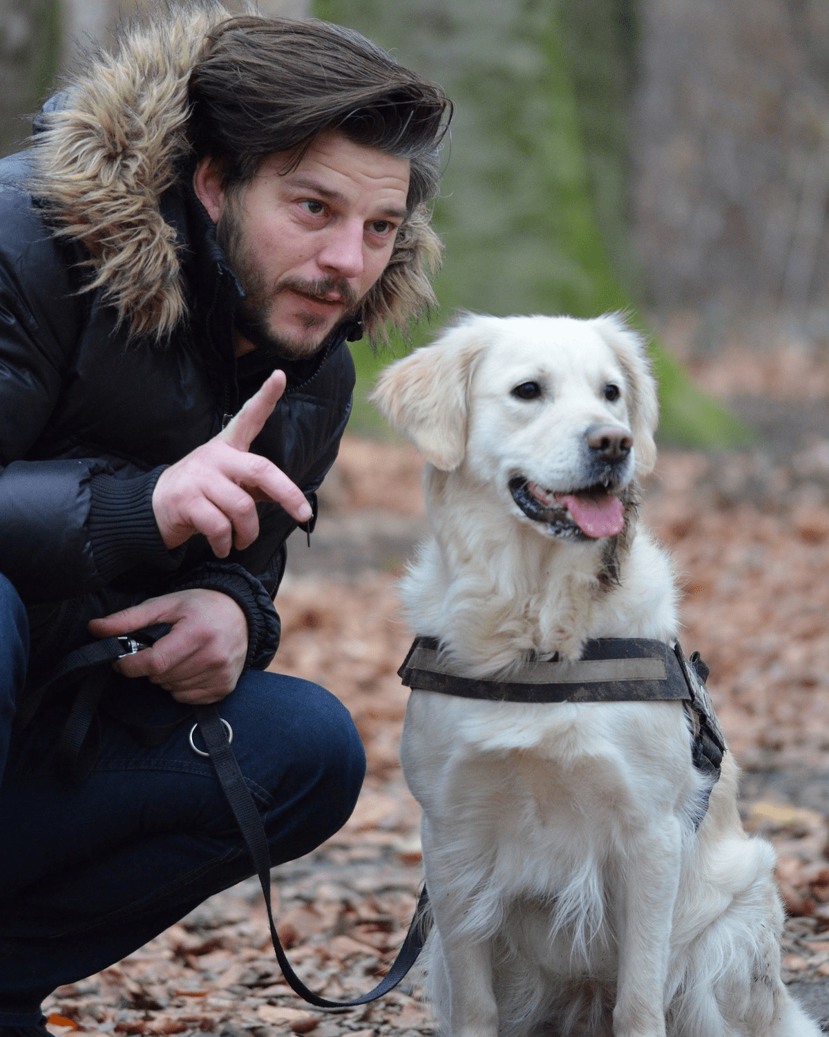 Ein Mann hockt neben seinem Hund im Freien und gibt ihm mit erhobenem Zeigefinger ein Zeichen