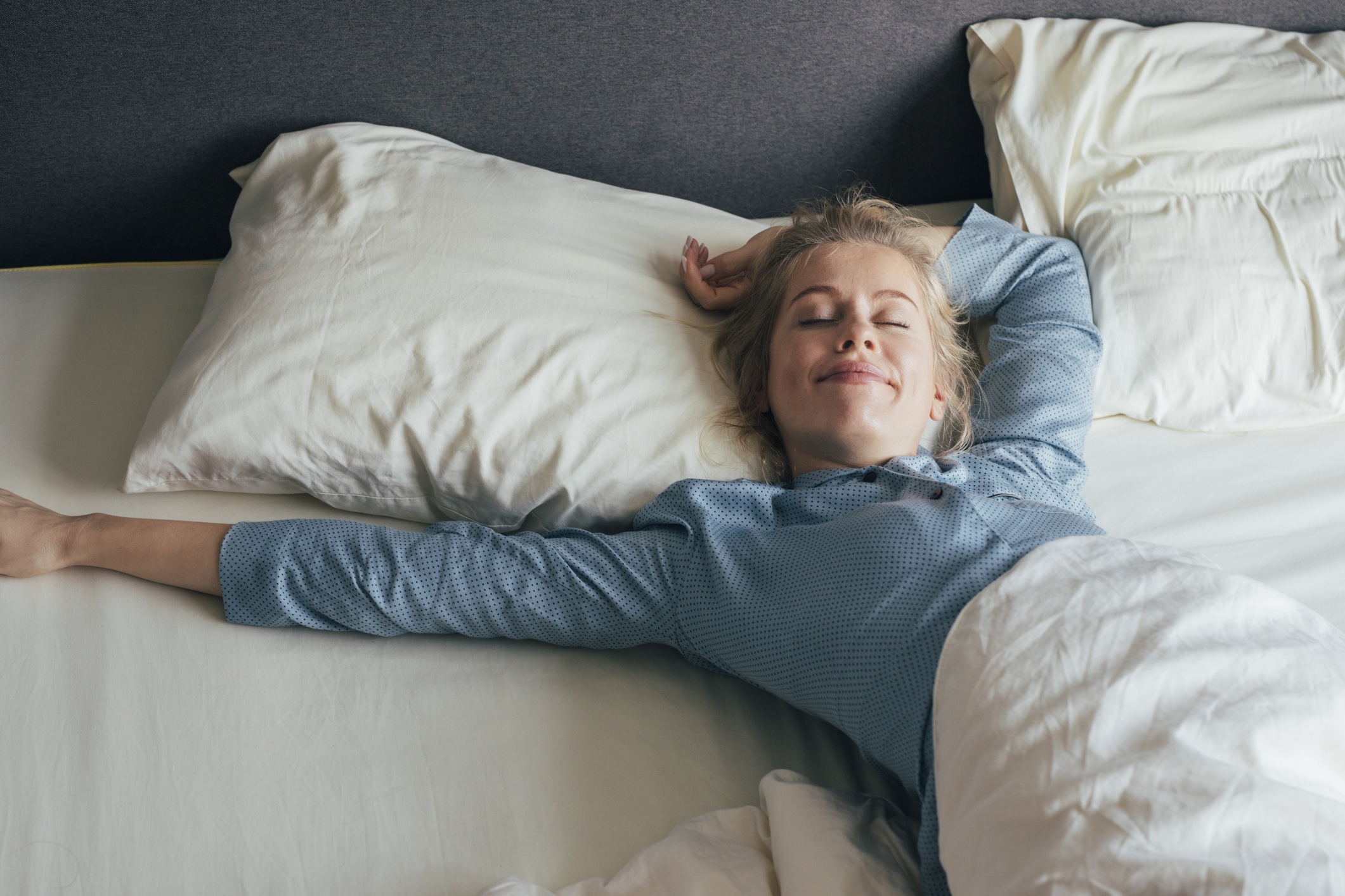 Eine junge Frau liegt mit zufriedenem Gesichtsausdruck und geschlossenen Augen auf einem Doppelbett und streckt die Arme von sich.