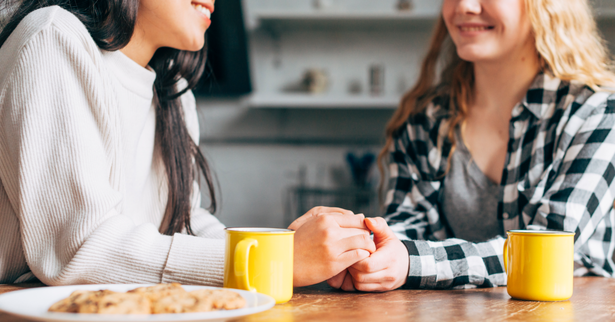 Studie zu Kaffee und PersÃ¶nlichkeit. Zwei Frauen halten sich lÃ¤chelnd die Hand, vor ihnen stehen zwei Kaffeetassen.