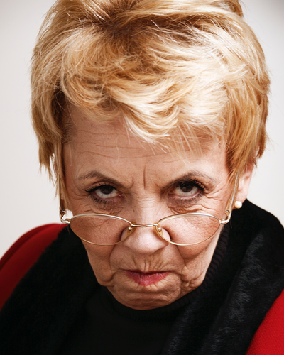 Eine ältere Dame mit kurzen blonden Haaren und einer Brille schaut grimmig in die Kamera.