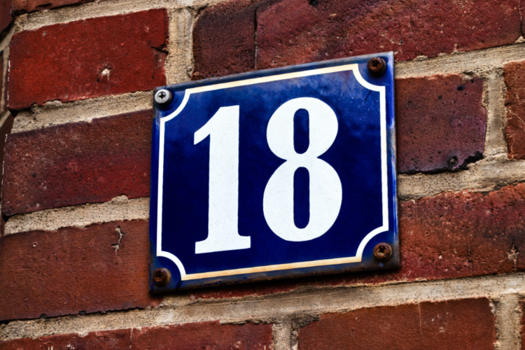 Ein altes Hausnummernschild mit der Nummer „18“. Die Hausnummer ist in Weiß auf blauem Untergrund gedruckt.