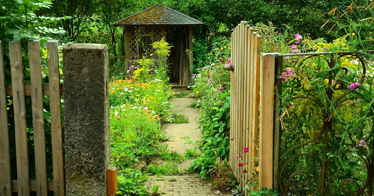 Ein offenes Tor in einem Holzzaun führt in einen Cottage Garden mit einer kleinen Holzhütte.