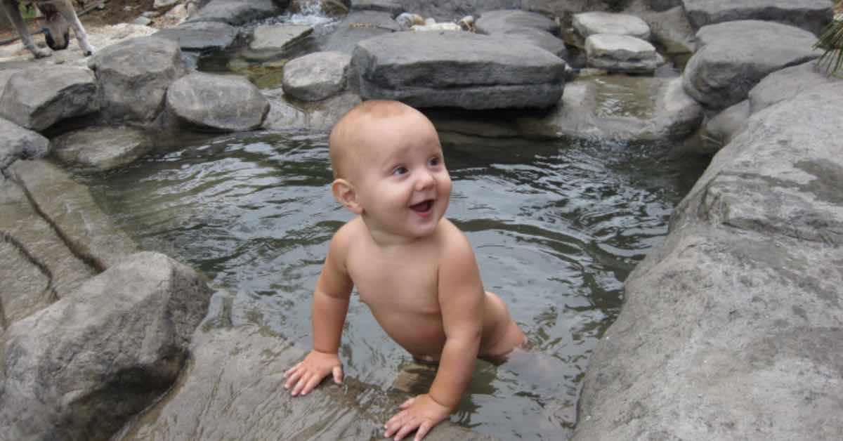 Kleinkind steht strahlend im Wasser. Der Rand des Pools sieht aus wie aus Felsgestein.