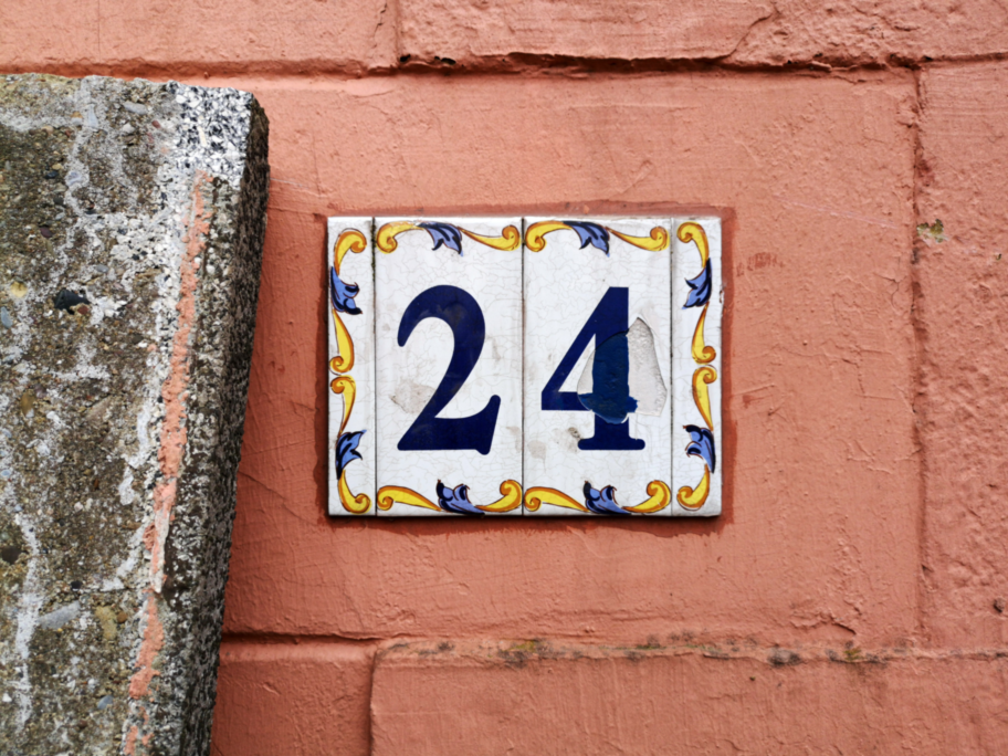 Ein altes Hausnummernschild mit der Nummer „24“. Das Schild ist leicht beschädigt und hängt an einer orangefarbenen Wand.