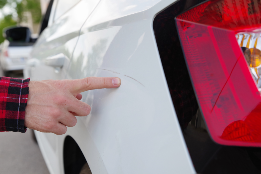 Jemand zeigt mit dem Zeigefinger auf einen Kratzer an einem weiÃŸen Auto. Mithilfe des KriechÃ¶ls WD-40 lassen sich leichte Lackkratzer entfernen.