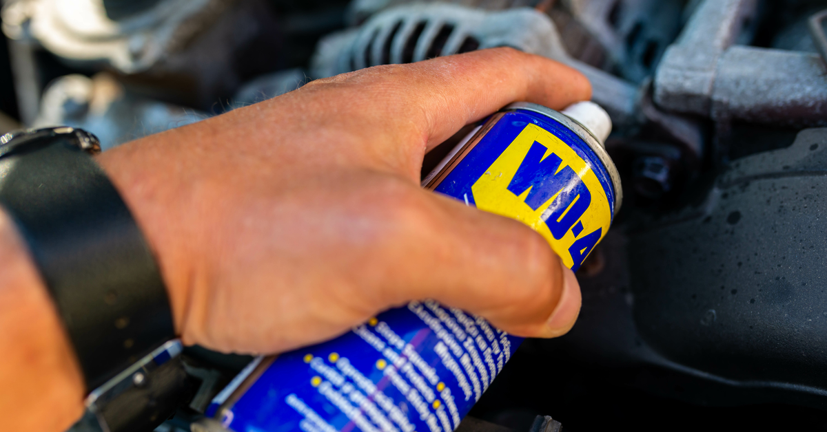 Detailaufnahme von einer Hand, die eine Dose WD-40 Ã¼ber Bauteile eines Motors hÃ¤lt und diese einsprÃ¼ht.