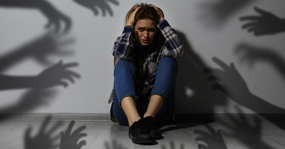 Eine Frau sitzt vor einer weißen Wand und hält sich die Ohren zu. Auf der weißen Wand sieht man Hände als Schatten.