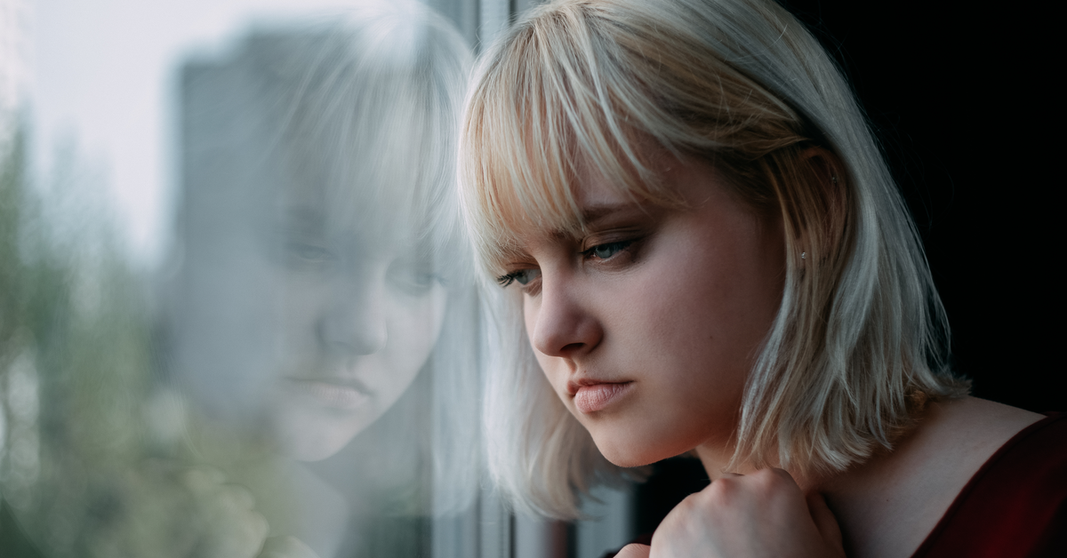 Eine junge, blonde Frau schaut traurig aus dem Fenster. Ihr Gesicht spiegelt sich in der Scheibe.