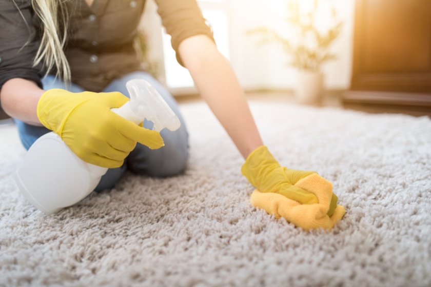 Eine Frau reinigt mit einem Spray und einem Mikrofasertuch einen Teppich. Sie hat gelbe Gummihandschuhe an.