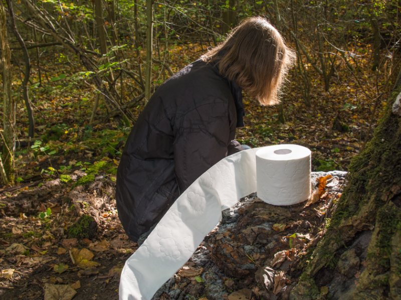 Eine Frau hockt neben einer Rolle Toilettenpapier in einem Wald.