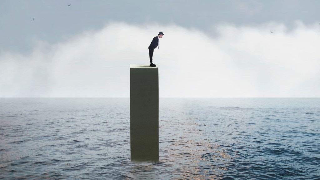 Mann steht auf einem Pfahl mitten im Meer und schaut in Wasser.