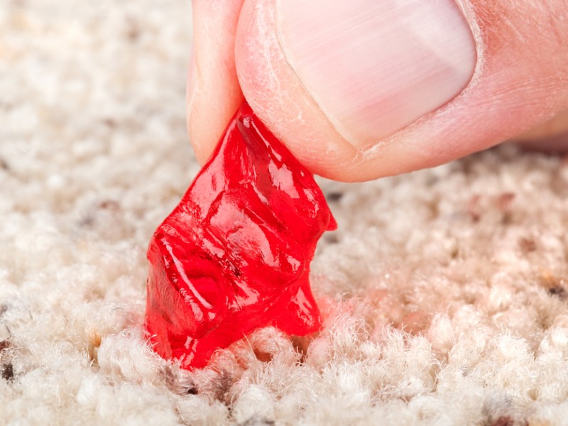 Ein roter Bonbon klebt in hellem Teppich fest.