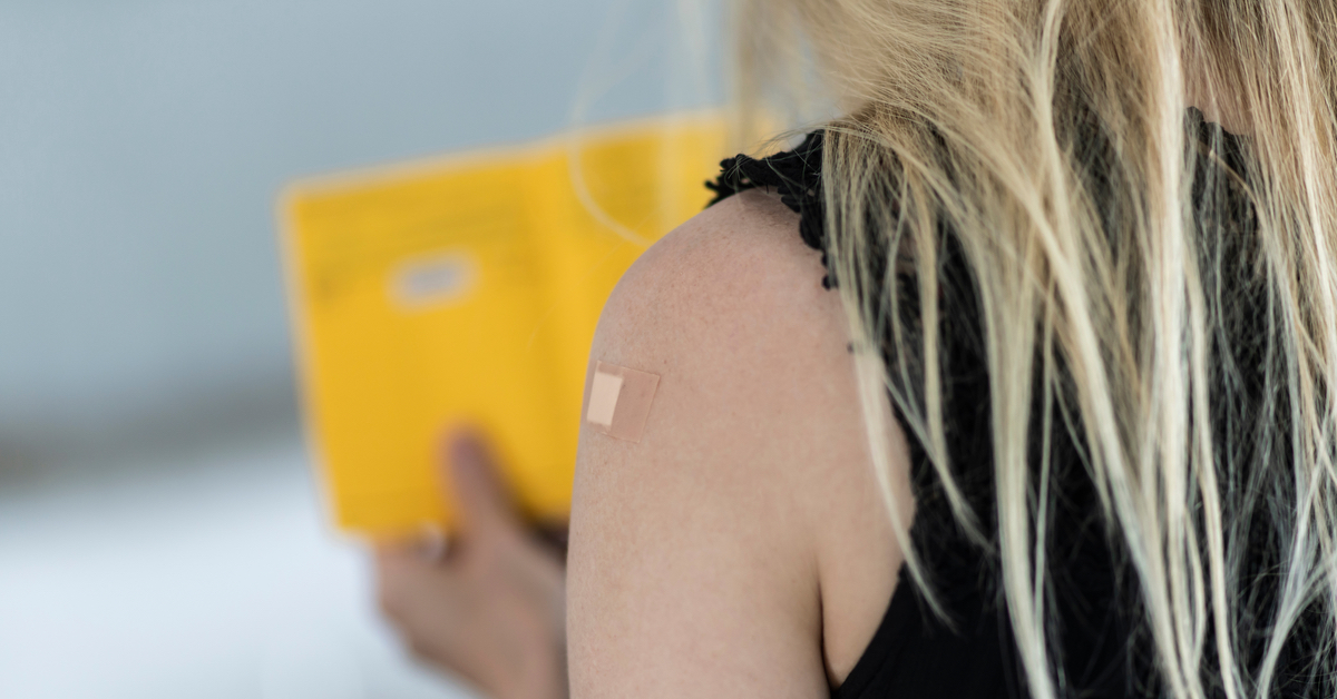 Eine blonde Frau von hinten fotografiert. Sie schaut sich ihren Impfpass an und man sieht ein Pflaster auf ihrem Arm.
