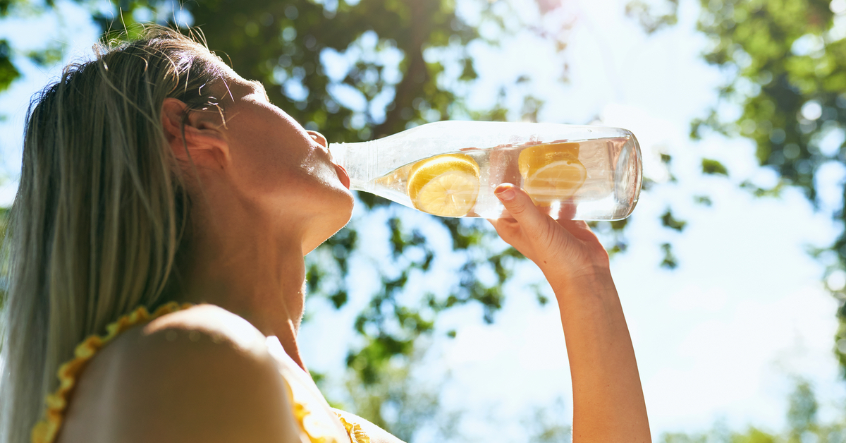 Eine Frau mit blonden Haaren in der Natur trinkt Zitronen-Wasser aus einer Flasche.