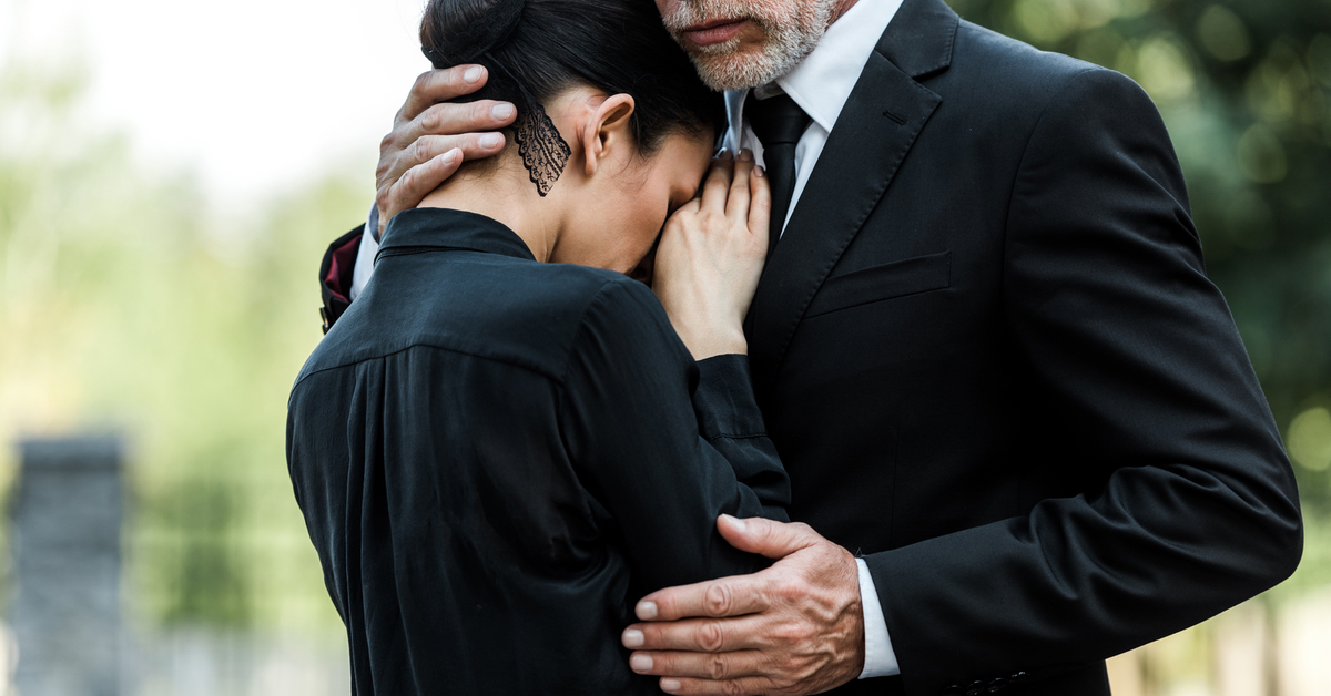 Eine Frau legt ihren Kopf auf die Brust eines Mannes, der sie mit einer Umarmung zu beruhigen scheint. Beide sind ganz in Schwarz gekleidet.