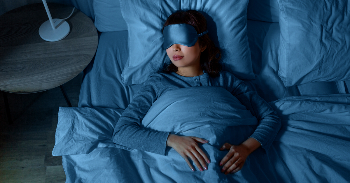 Eine Frau liegt im Bett mit blauer BettwÃ¤sche und trÃ¤gt eine Schlafbrille.