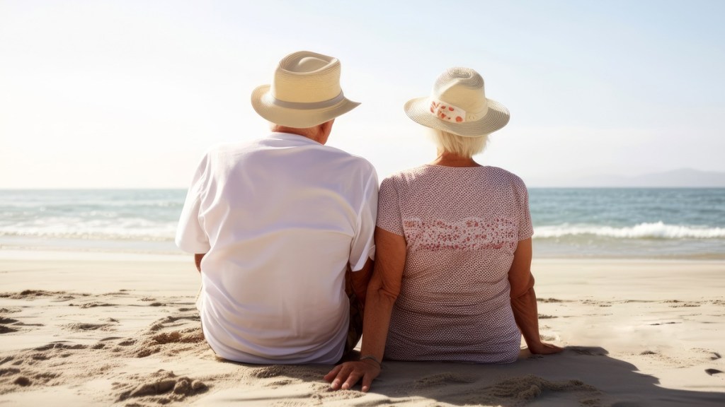 Ein älteres Paar sitzt verliebt am Strand und schaut gemeinsam aufs Meer. 