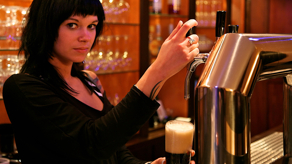 Eine schwarzhaarige Barfrau zapft ein Bier und schaut in die Kamera.