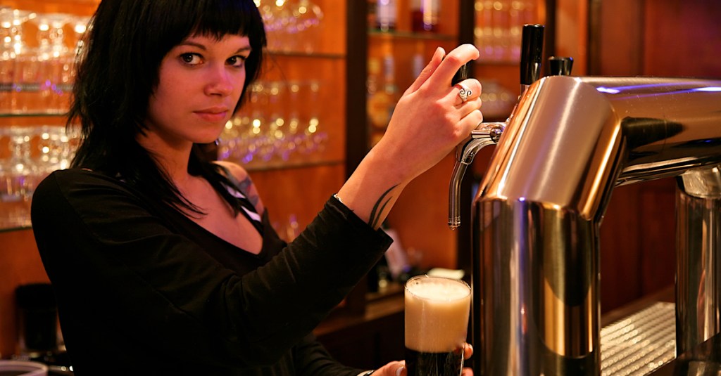 Eine schwarzhaarige Barfrau zapft ein Bier und schaut in die Kamera.