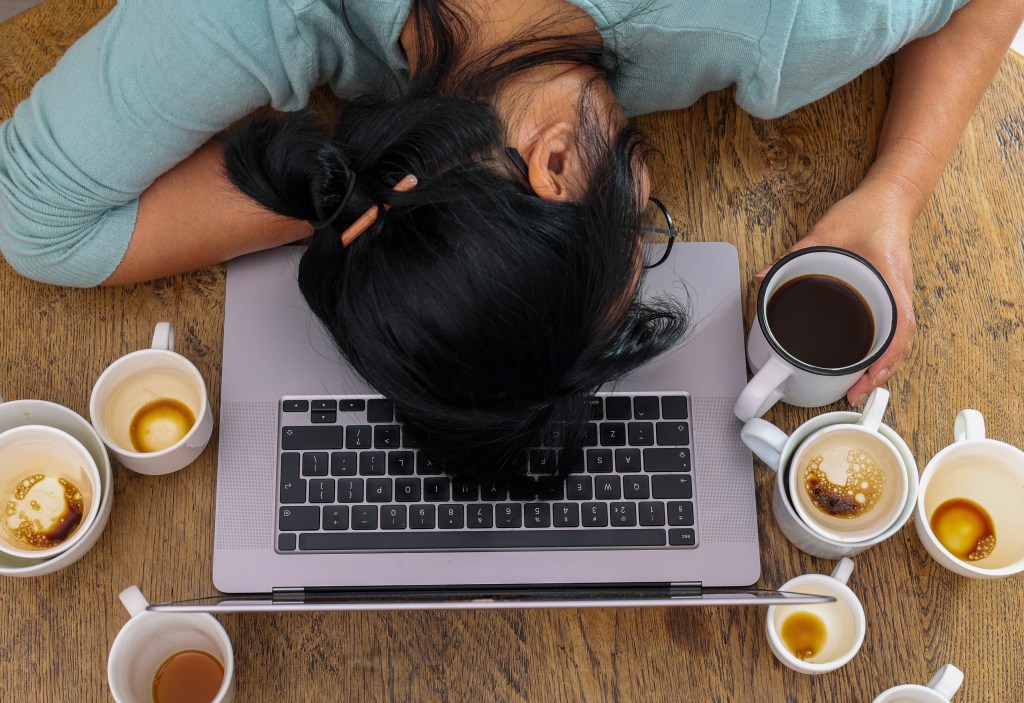 Eine Frau bricht über ihrem Laptop zusammen und versucht ihre Müdigkeit mit Kaffee zu kompensieren.