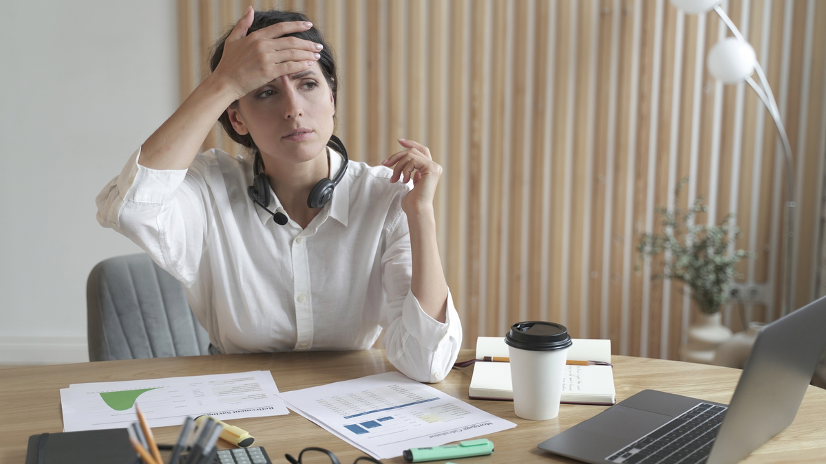 Eine Frau sitzt am Schreibtisch und greift sich an den Kopf, als habe sie Kopfschmerzen.
