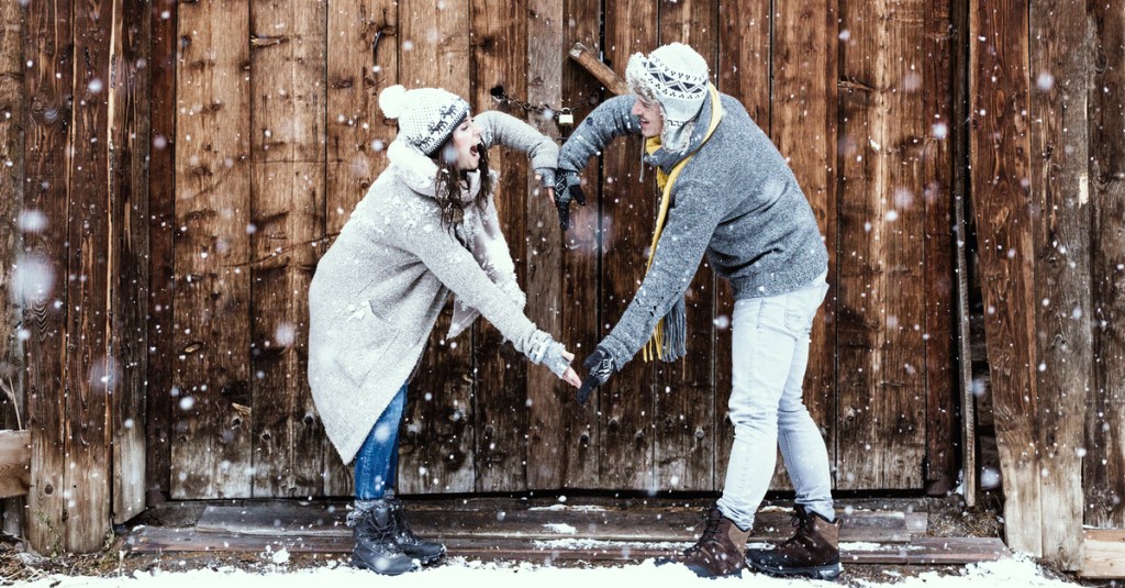Ein Pärchen, das im Schnee vor einer Holzwand ein Herz mit seinen Armen formt.