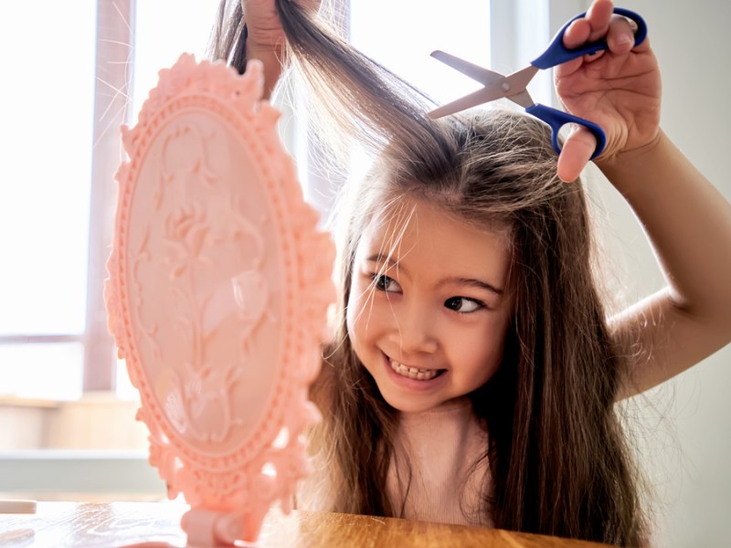 Ein Mädchen versucht vor einem Handspiegel ihre Haare zu schneiden.