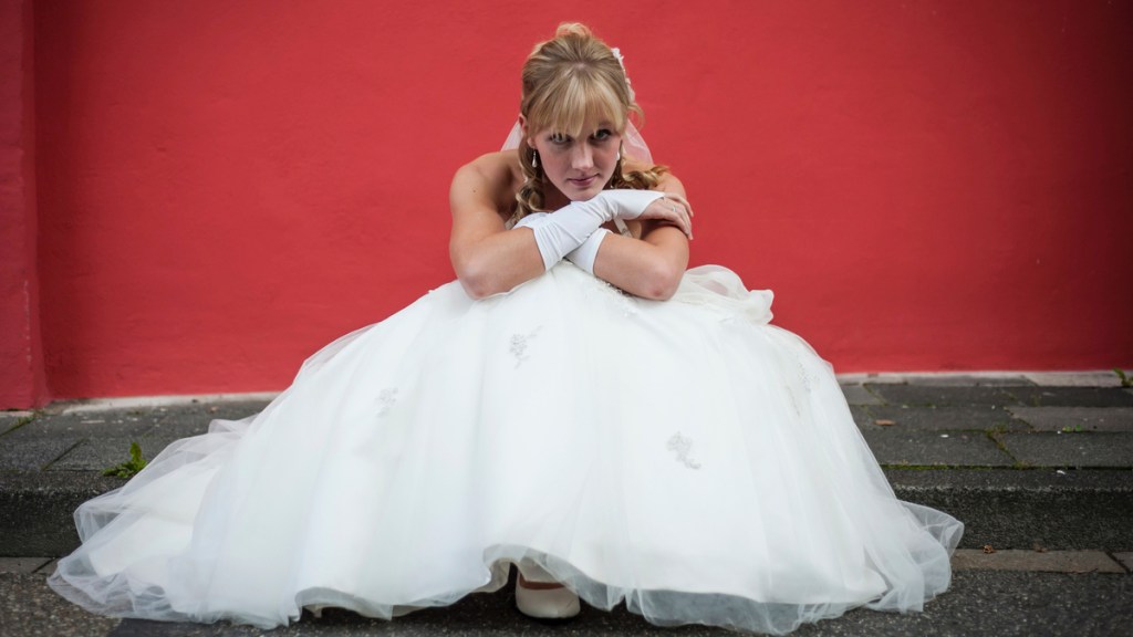Eine blonde Frau im Hochzeitskleid sitzt vor einer roten Wand auf dem Bürgersteig und stützt ihr Kinn traurig auf ihre Knie.